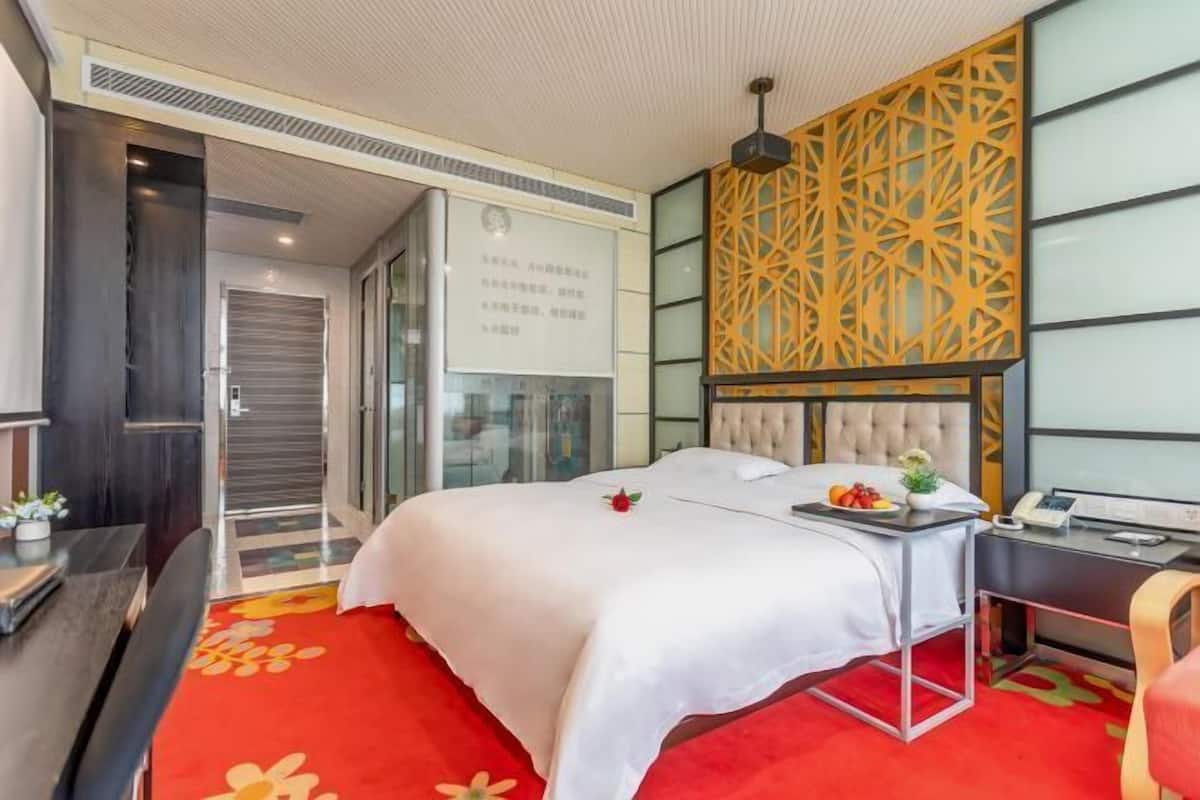 A hotel room with a bed, a desk, a chair, a small table with fruits, and a decorative wall panel.