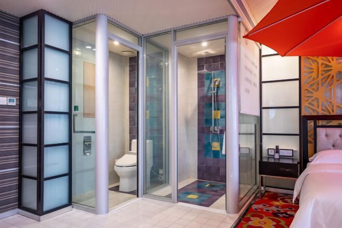 A modern hotel room with a glass-enclosed shower, a toilet, and a bed with a red umbrella.