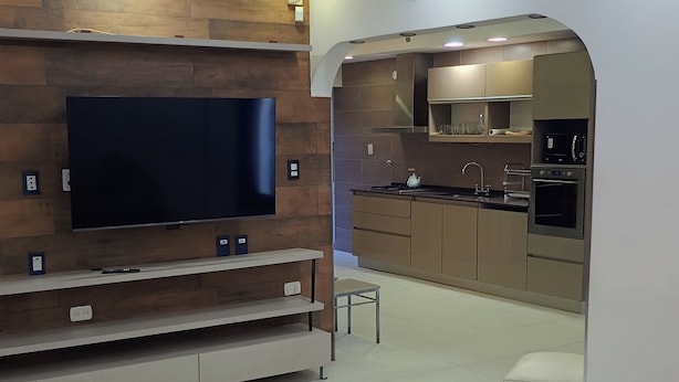 Un salon moderne doté d’un téléviseur à écran plat, d’un canapé et d’un coin cuisine visible à travers une arche.