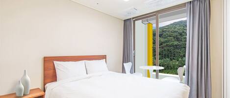 Habitación de hotel con una cama grande, una mesita de noche con dos jarrones, una pequeña mesa redonda y vistas a zonas verdes a través de una ventana con cortinas.