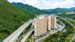 A multi-story building with a modern design, surrounded by lush green hills and a river. - Ariari Hotel (Jeongseon)