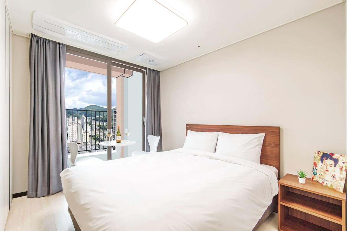 Ett hotellrum med en stor säng, ett litet bord med en stol, en balkong med gardiner och utsikt över staden.
