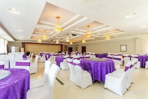 Un salón de banquetes con mesas redondas cubiertas con manteles blancos y centros de mesa morados, dispuestas en filas.
