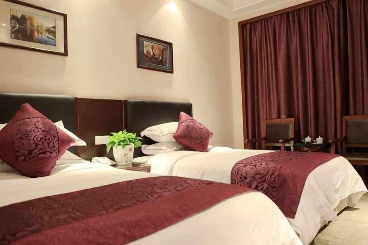Habitación de hotel con dos camas, una mesita de noche, una planta en maceta y un ventanal con cortinas.