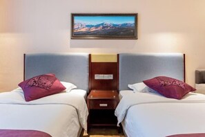 A hotel room with two beds, each with a wooden nightstand and a small table. A framed landscape painting is on the wall above the beds.