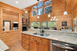 Une cuisine dotée de plans de travail en granit, d’un lave-vaisselle, de meubles en bois, d’une cheminée en pierre et de grandes fenêtres.