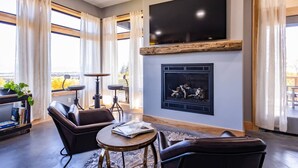 A modern living room with a fireplace, leather sofas, a coffee table, and a large TV mounted on the wall. - Lakeshore Lodging Inlet View Home (Homer)