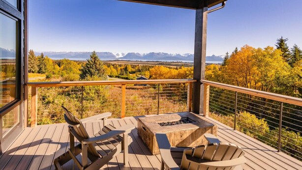 A wooden deck with outdoor seating and a fire pit, overlooking a mountainous landscape with autumn foliage. - Lakeshore Lodging Inlet View Home (Homer)
