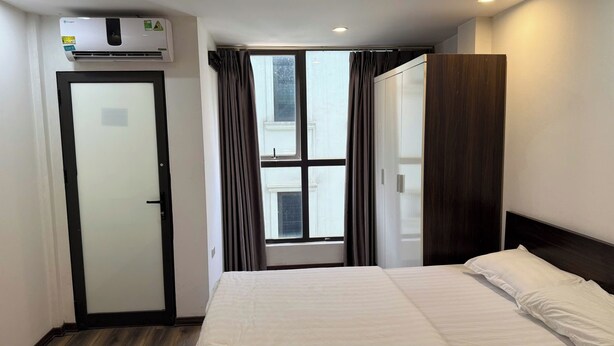 A hotel room with a bed, a wardrobe, a window with curtains, and an air conditioning unit. - Era Apartment Ha Yen Quyet (Hanoi)