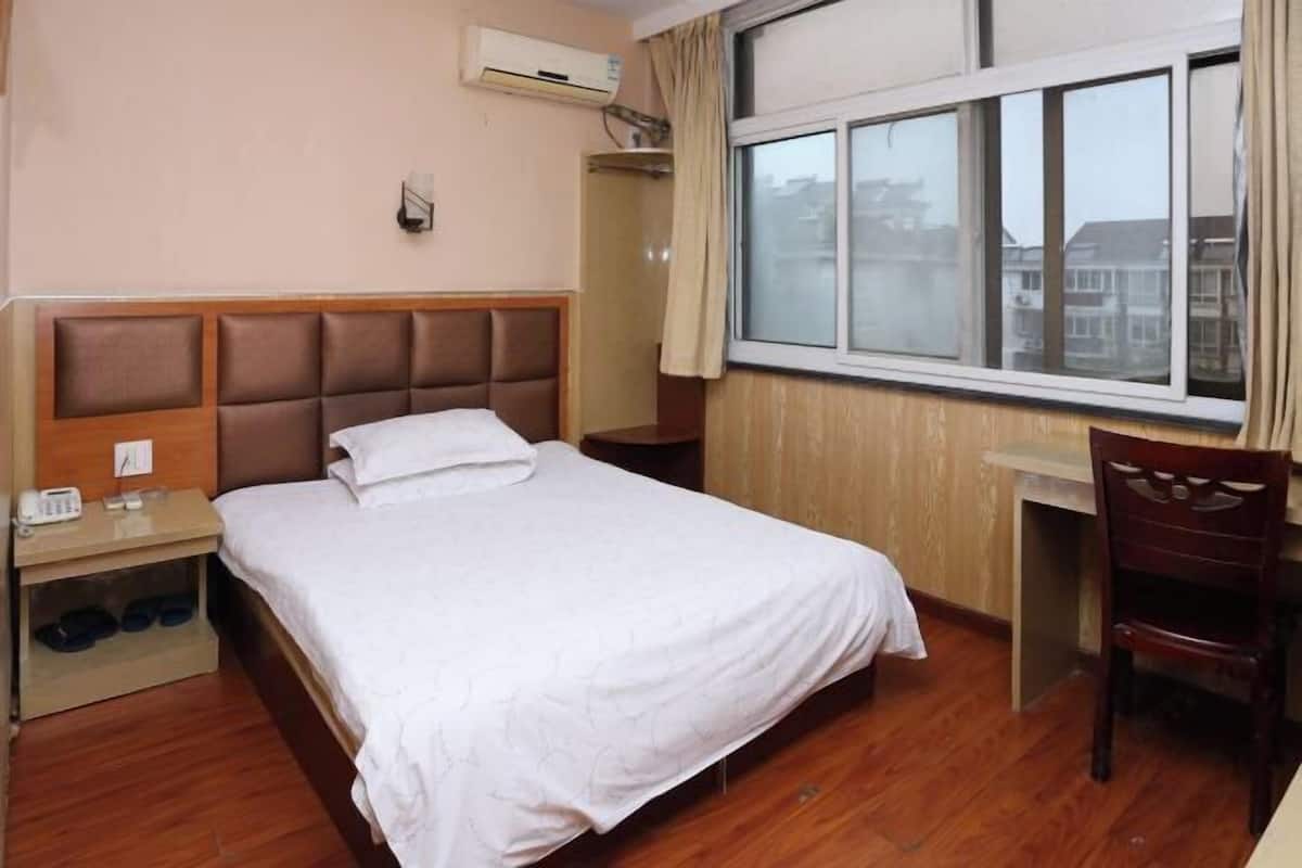 A hotel room with a bed, a desk, a chair, a wardrobe, and a window with a view of buildings.
