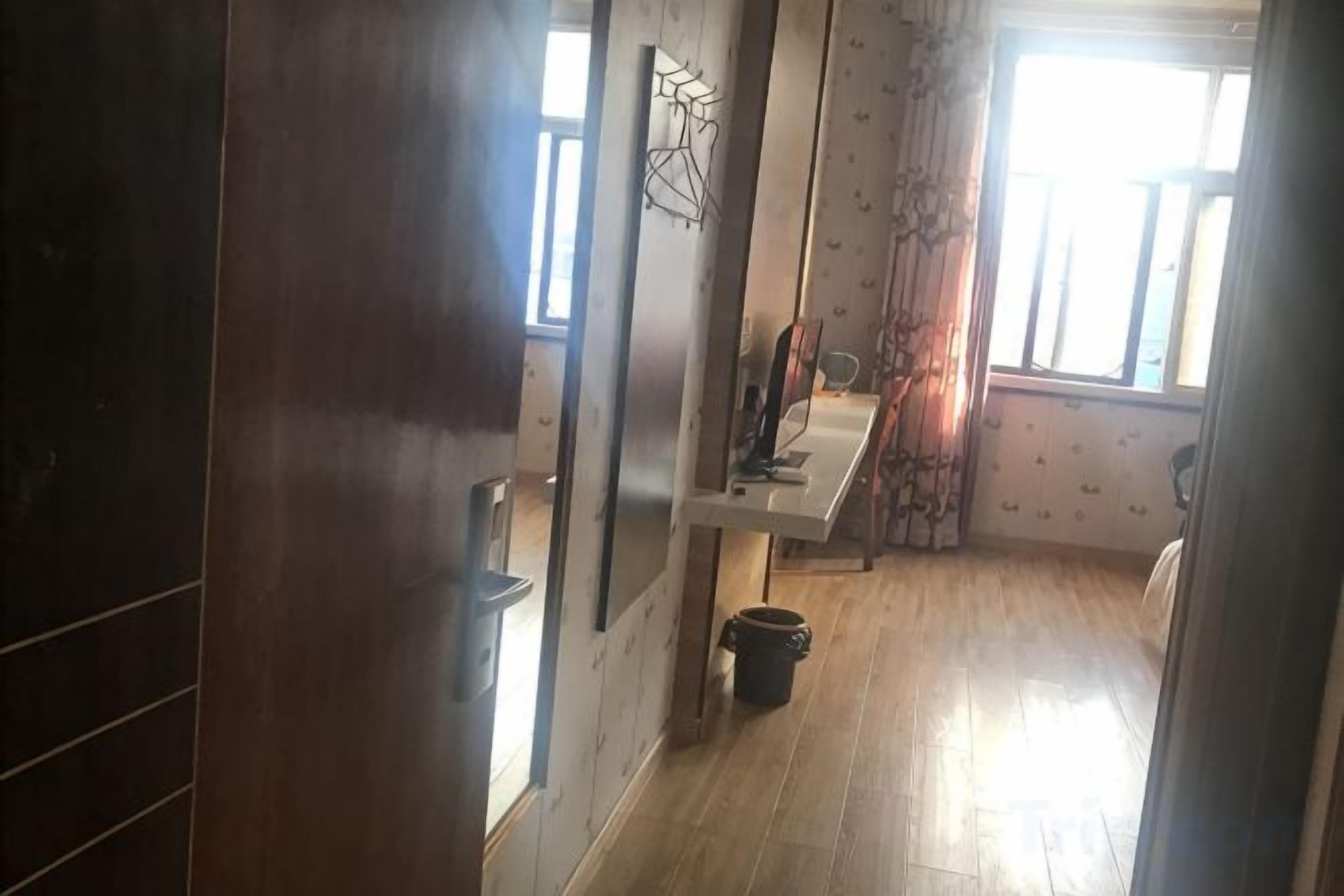 A narrow hallway with a wooden floor leading to a room with a window and a curtain.