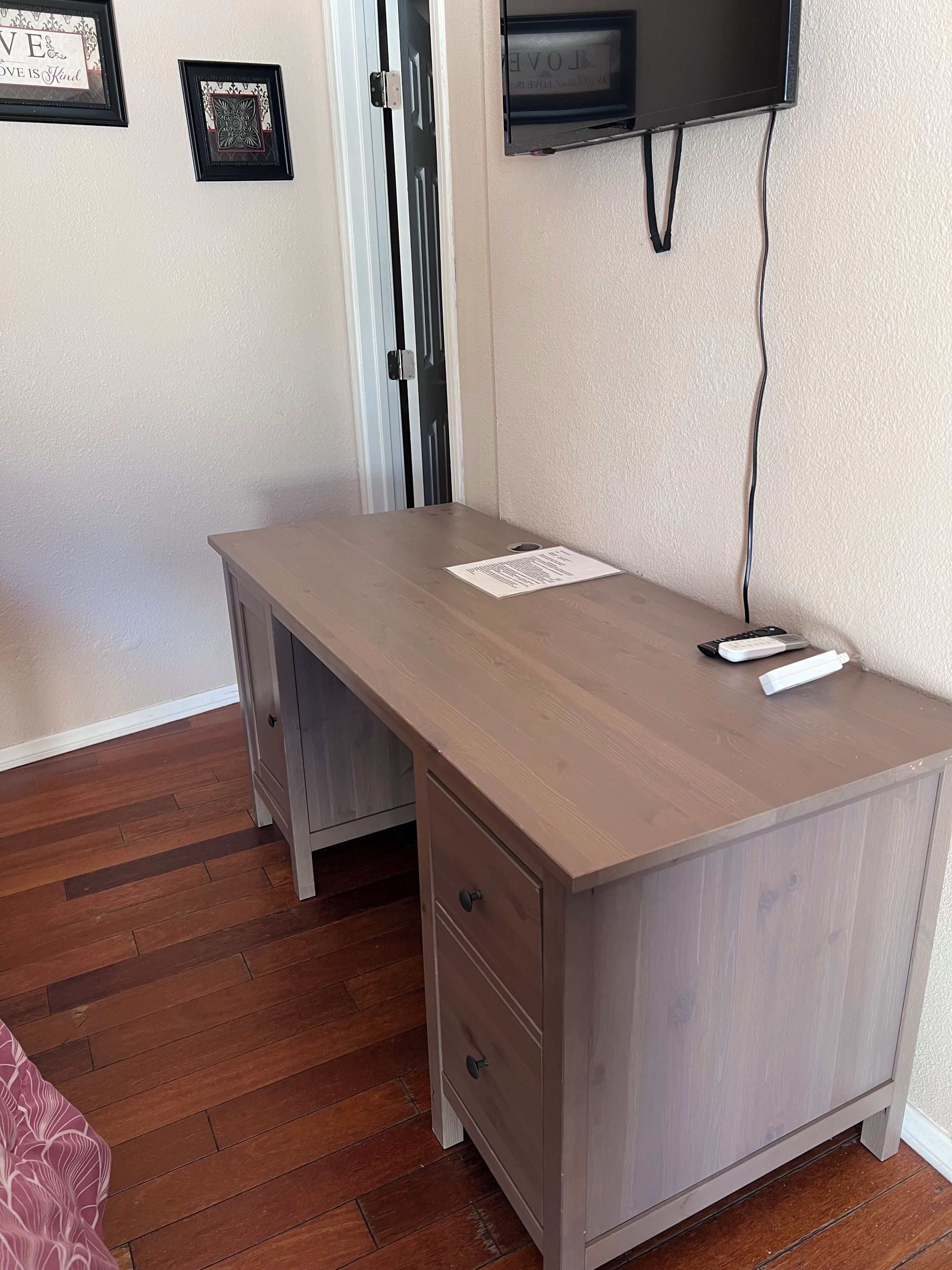 Quarto com escrivaninha de madeira, televisão fixada na parede e porta com puxador de metal.