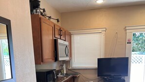 A compact kitchenette with a microwave, coffee maker, and a small refrigerator. There is a wooden table with a plant, a TV on a stand, and a window with blinds. - Charming  Bedroom. In home (Riverside)