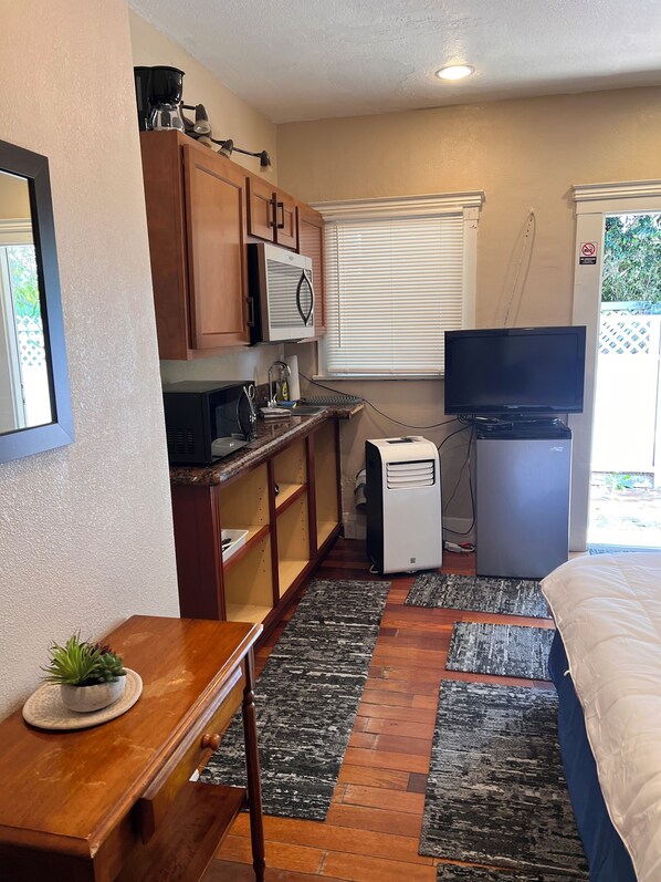 A compact kitchenette with a microwave, coffee maker, and a small refrigerator. There is a wooden table with a plant, a TV on a stand, and a window with blinds.