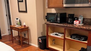 A compact hotel room with a wooden floor, a bed with white bedding, a small table with a plant, a microwave, a coffee maker, and a TV stand with a remote control. - Charming  Bedroom. In home (Riverside)