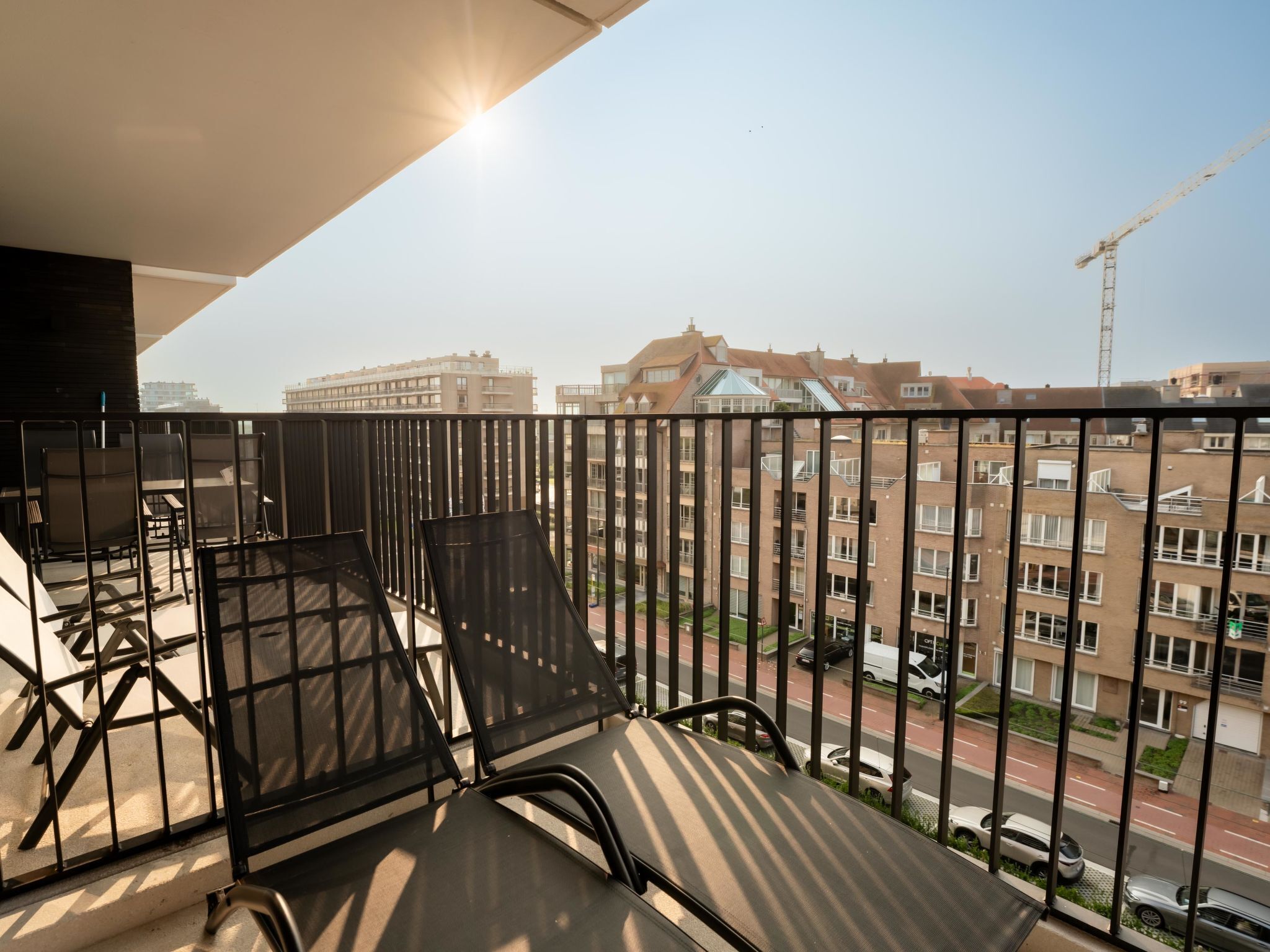 Un balcone con ringhiere in metallo, due poltrone nere e vista sulla città con edifici e una gru edile.