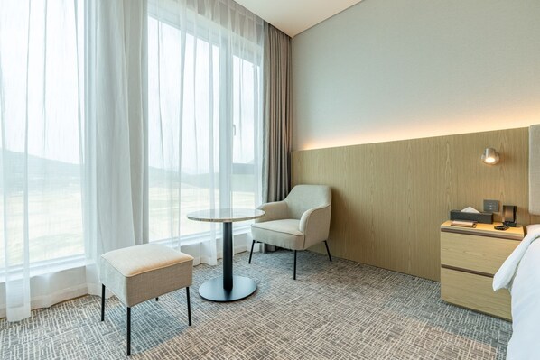 Habitación de hotel con un ventanal grande, una mesa redonda, una silla, un pequeño otoman, una mesita de noche con lámpara y una pared de acento de madera.