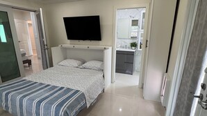 Une chambre à coucher avec un lit, un téléviseur fixé au mur et une salle de bain visible à travers une porte ouverte.