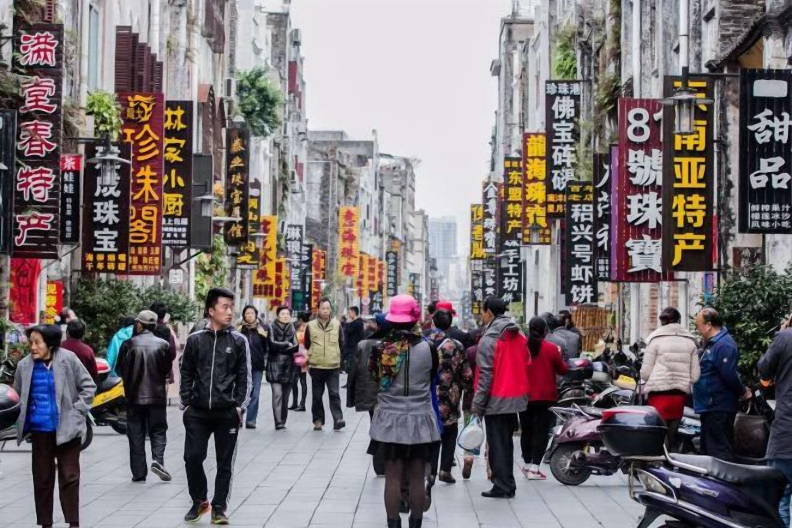 A busy street with pedestrians and motorcycles, surrounded by buildings with large Chinese characters.