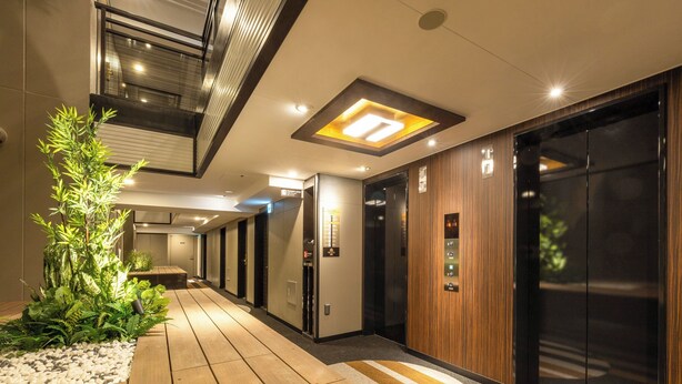 A modern hallway with wooden flooring, a long table, potted plants, and elevators. - APA Hotel Tokyo Nishikasai Ekimae (Tokyo)