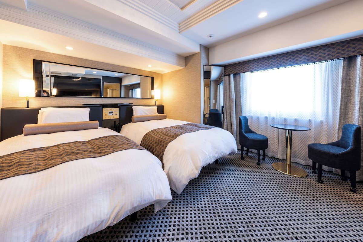 A hotel room with two beds, a small table, two chairs, a TV, and a large window with curtains.