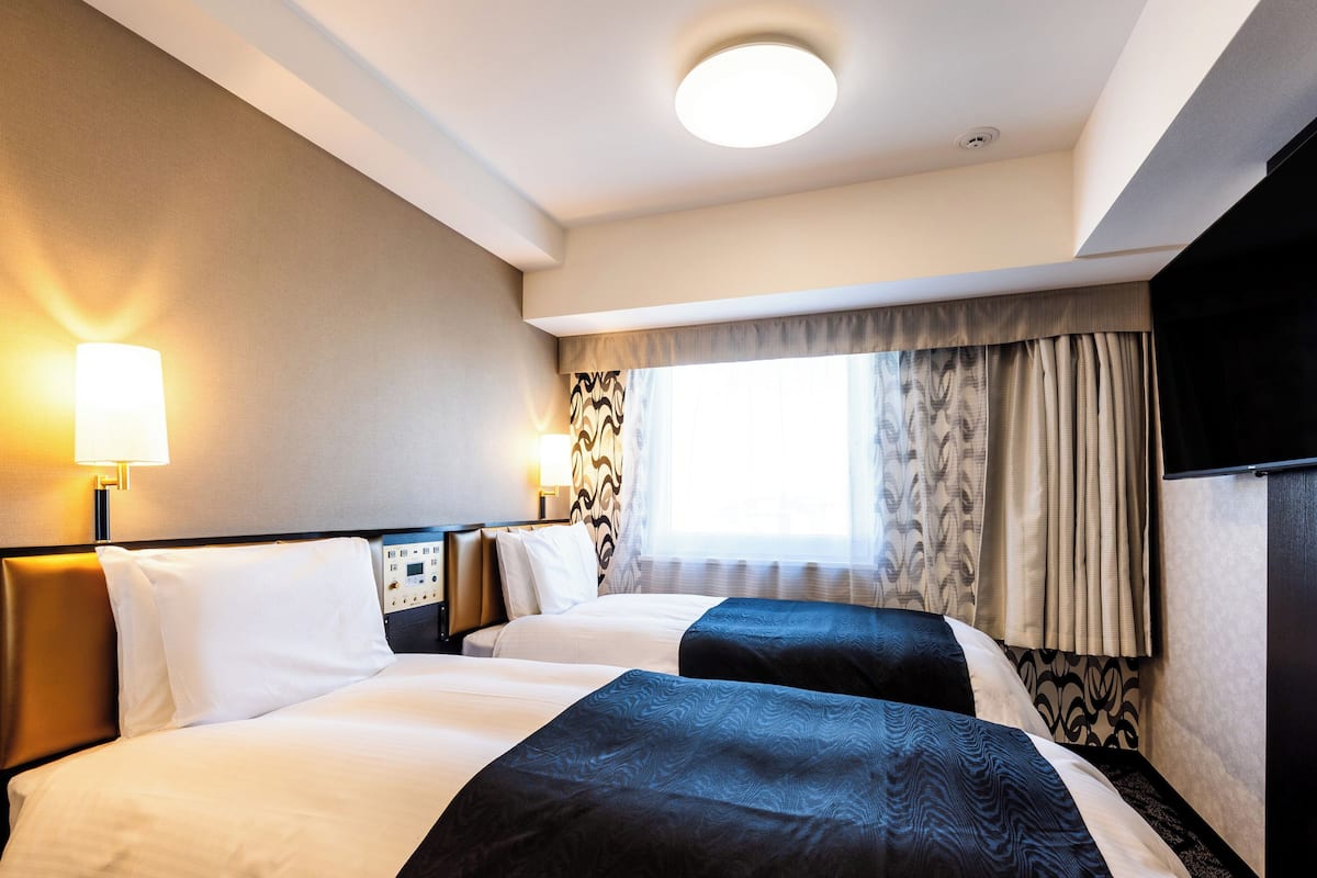 A hotel room with two beds, a control panel, a TV, and a window with curtains.