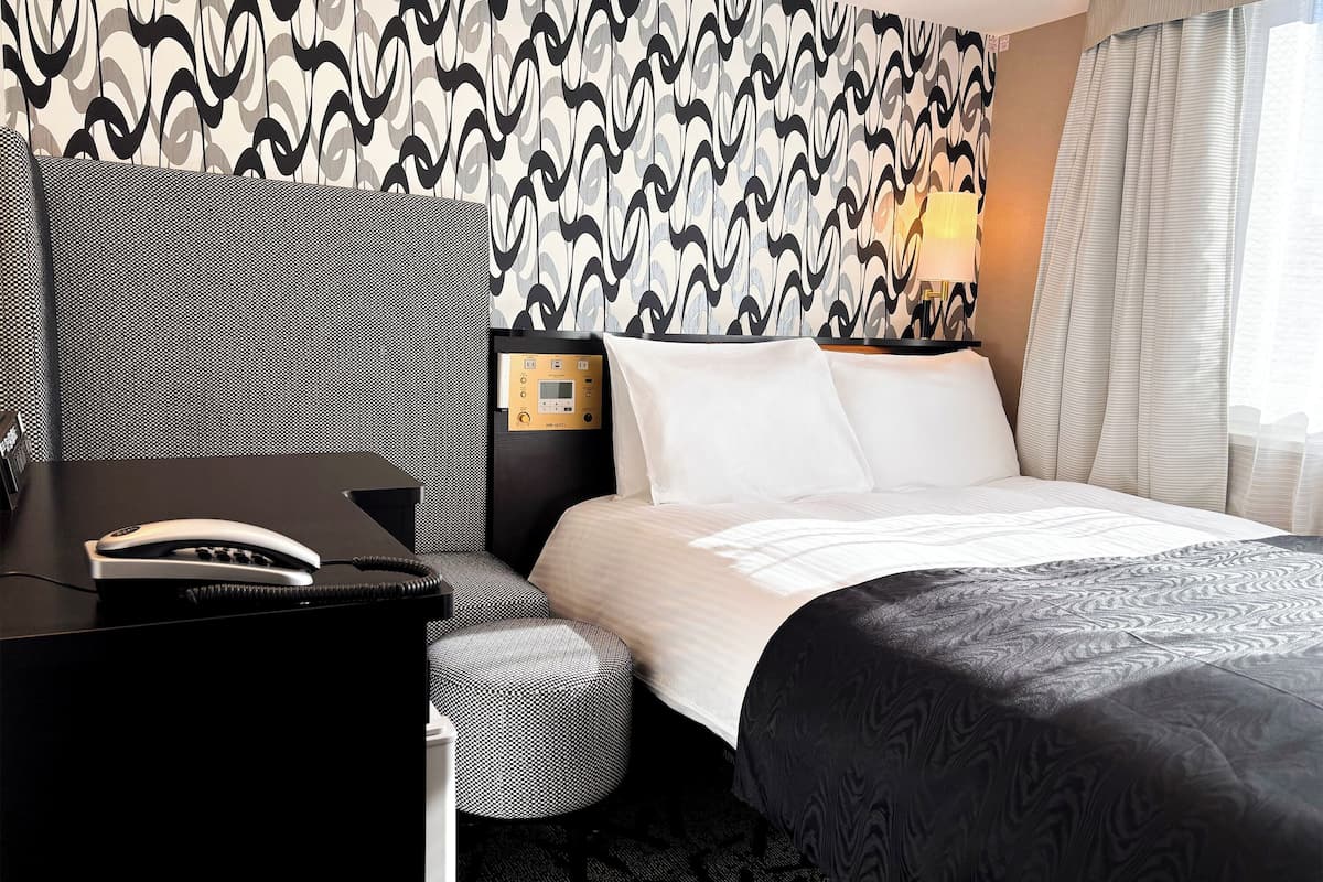A hotel room with a patterned wallpaper, a bed with white linens, a desk with a telephone, and a bedside table with a lamp.