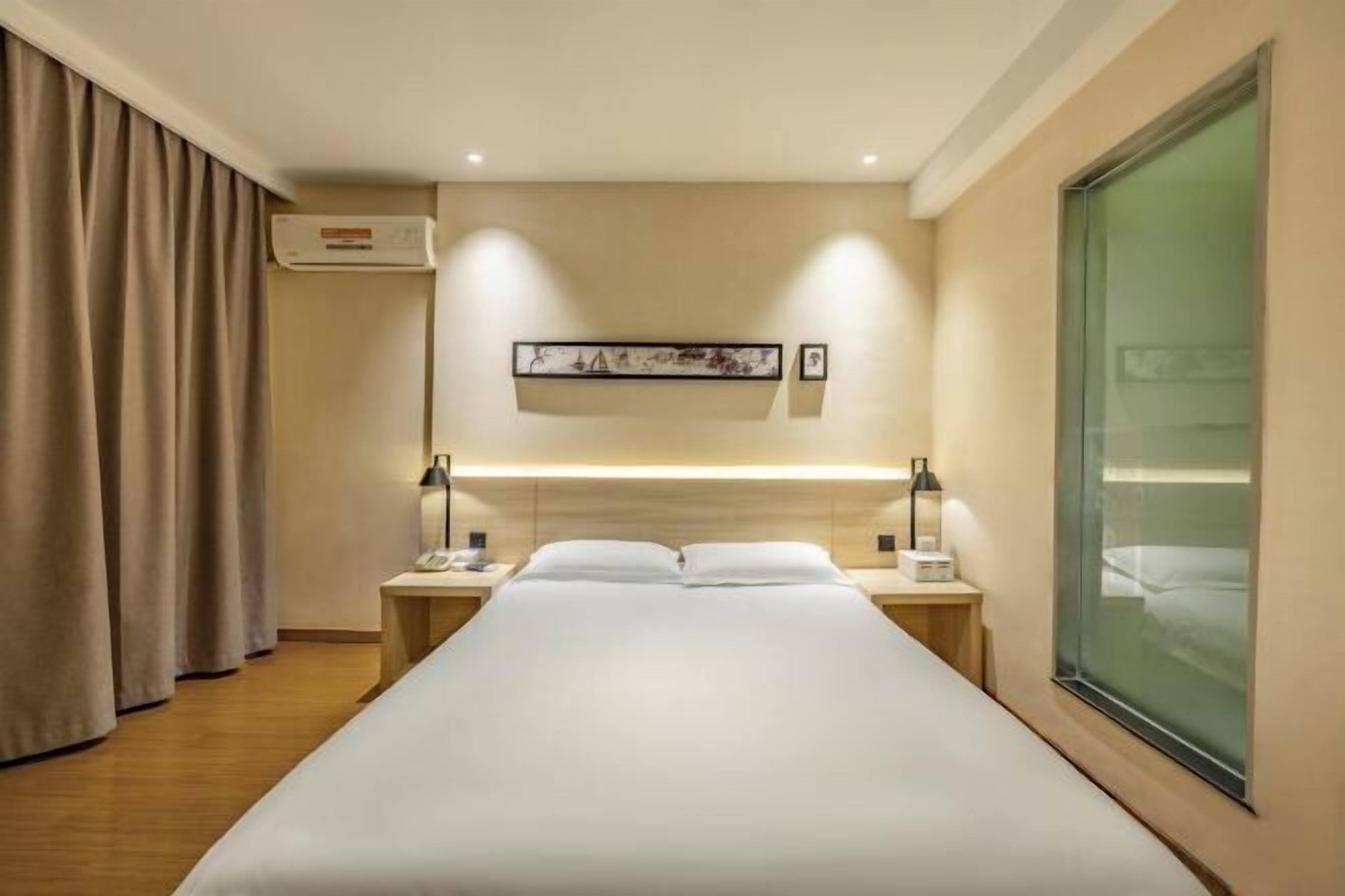 A hotel room with a bed, bedside tables, a wall-mounted air conditioner, a framed artwork, and a large window.