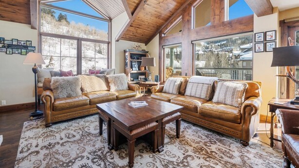 A spacious living room with high ceilings and large windows, featuring leather sofas, a wooden coffee table, and a patterned rug.
