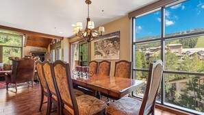 A dining area with a large wooden table and patterned chairs, a chandelier, and a view of a mountainous landscape through large windows.