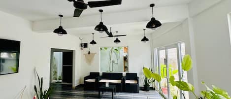 Un salón amplio con varios ventiladores de techo, un conjunto de sofá negro, una mesa de centro de madera y diversas plantas en macetas.