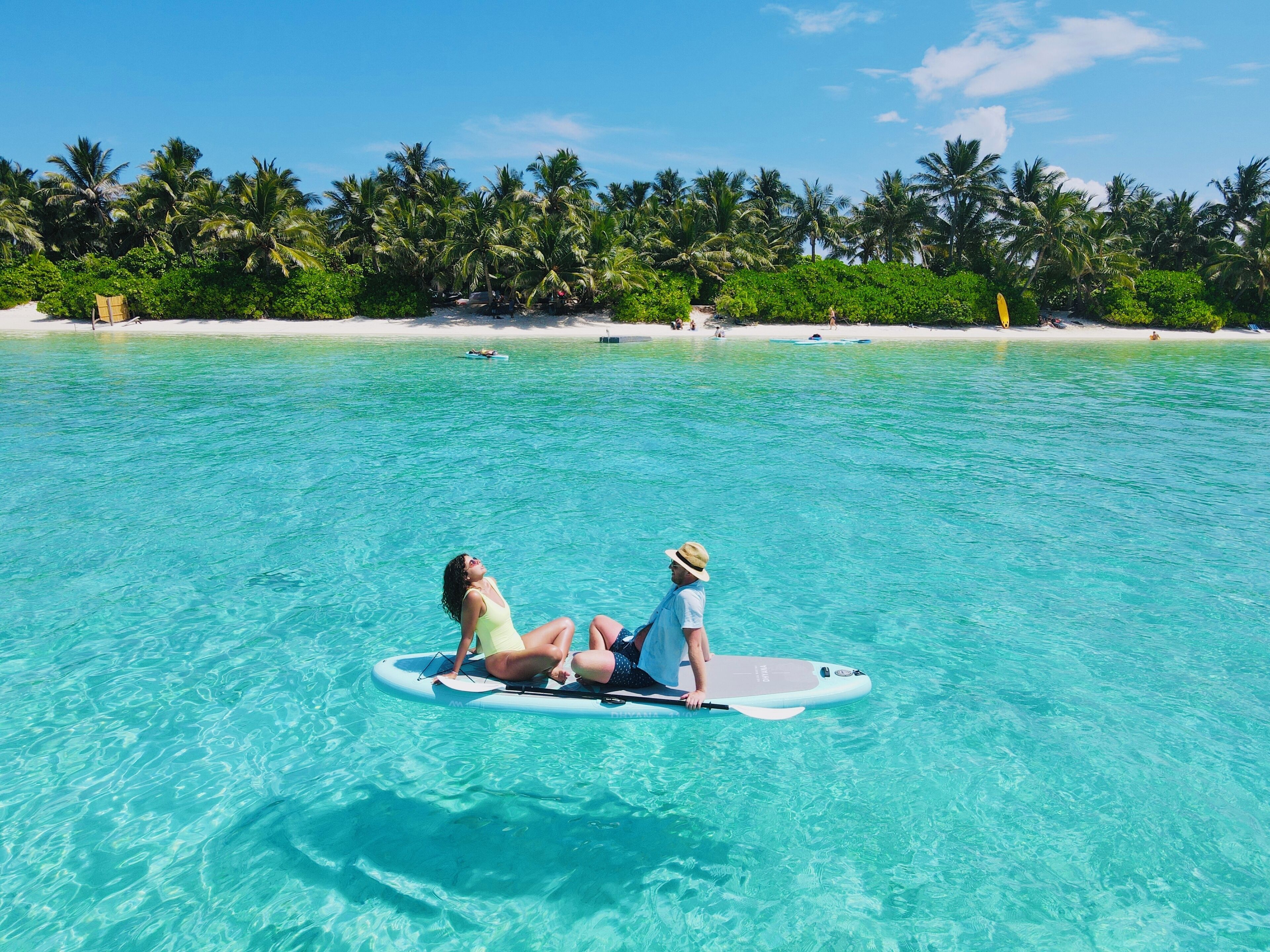 Two people on a paddleboard in clear turquoise water with a beach and palm trees in the background.