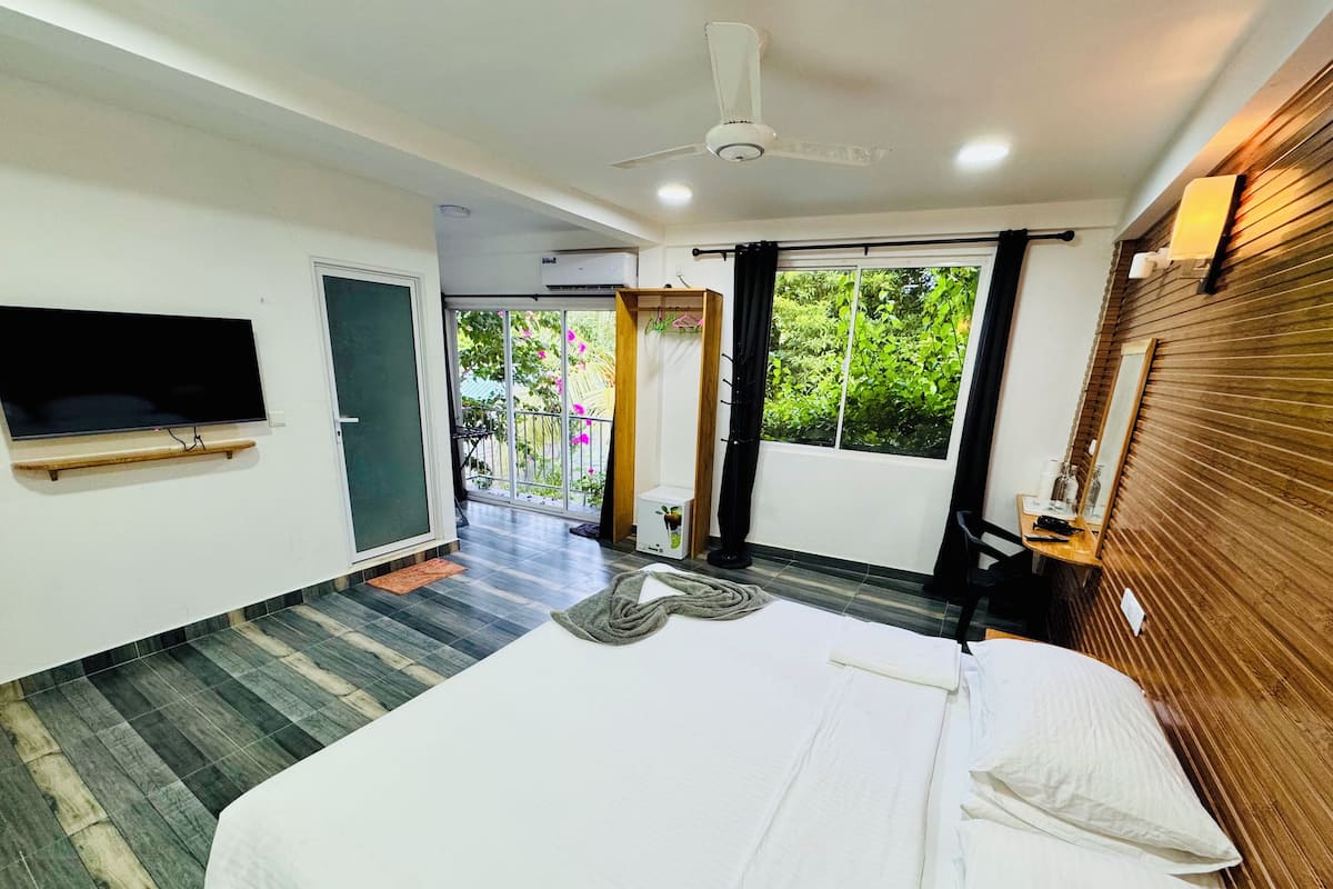 Ein modernes Hotelzimmer mit einem großen Bett, einem flachbildfernseher an der Wand und einem Balkon mit Blick auf Grünflächen.