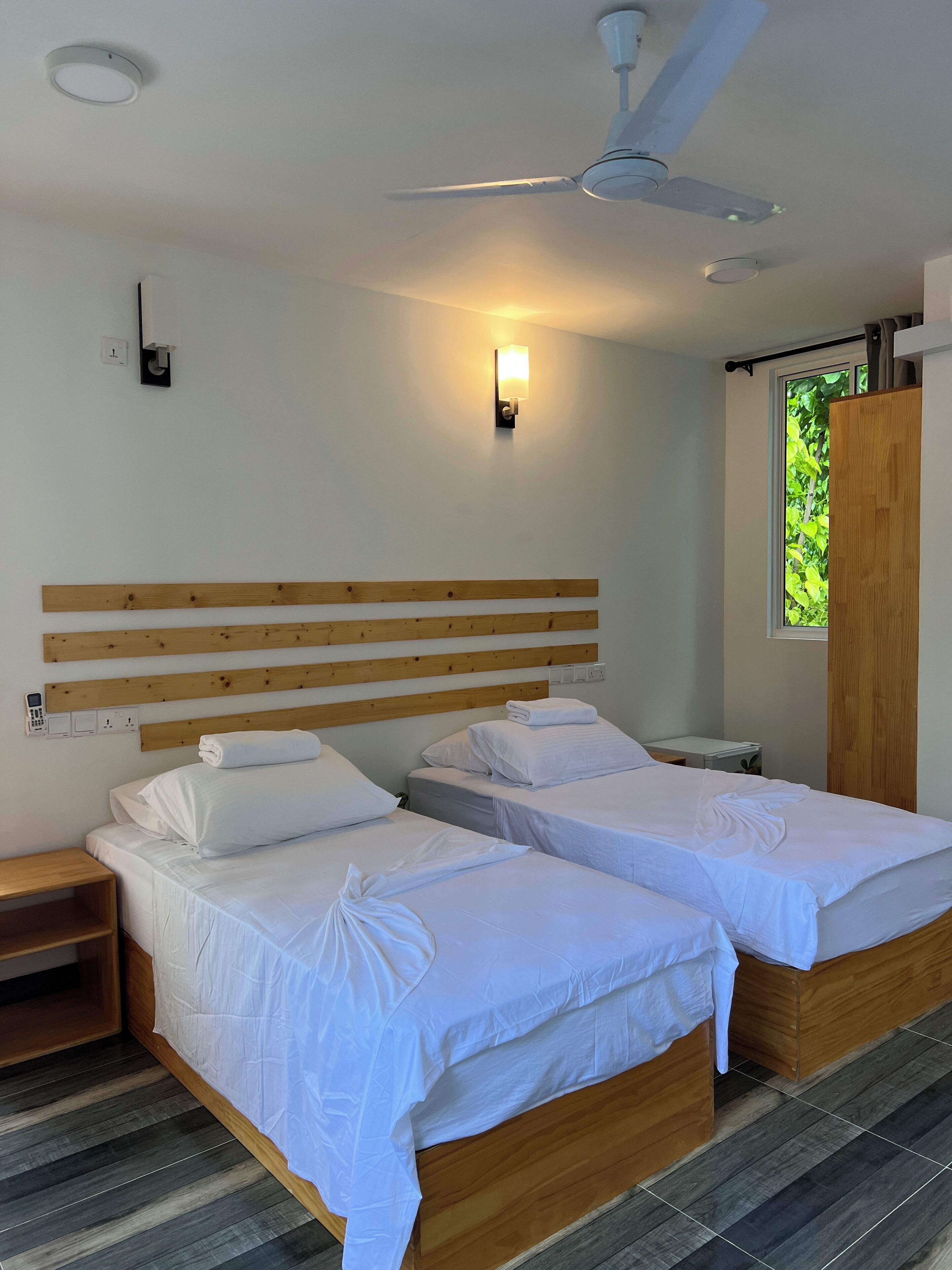 Hotellihuone, jossa on kaksi sänkyä, kattotuuletin, seinään kiinnitetty valaisin, ikkuna, josta on näkymä vehreään maisemaan, ja puinen sängynpääty.