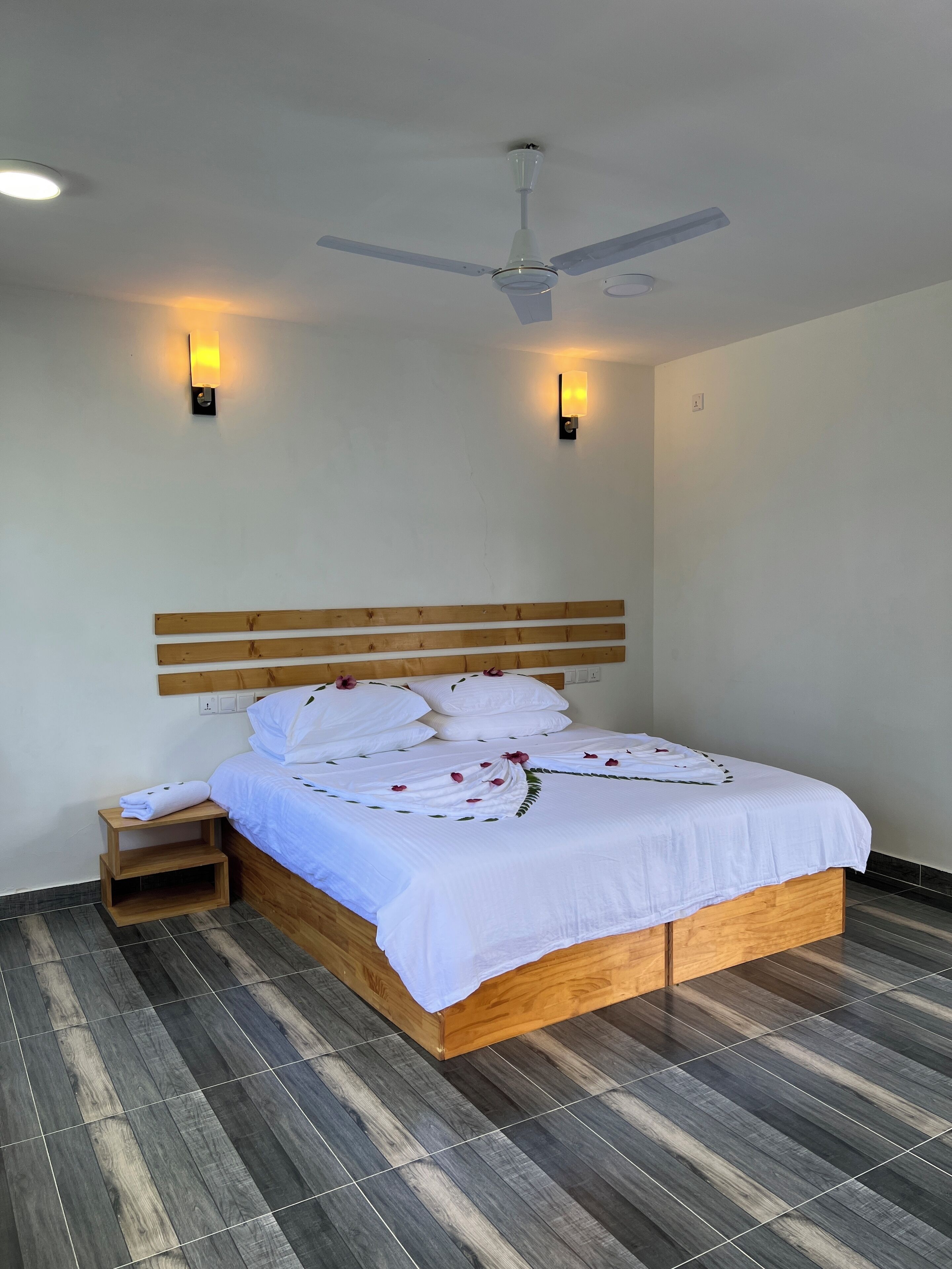 Un lit bien fait, avec du linge de lit blanc et une tête de lit en bois, une petite table de chevet en bois et un ventilateur de plafond.