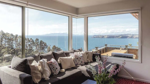 A modern living room with a large window overlooking a scenic view of the sea and mountains. - Seaview Sanctuary - Our Island (Waiheke Island)