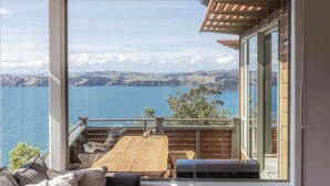 A balcony with a wooden table and chairs, overlooking a lake with distant hills. - Seaview Sanctuary - Our Island (Waiheke Island)