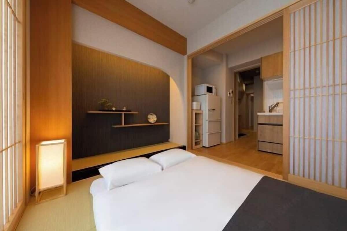 Habitació d'hotel moderna amb un llit gran, una tauleta de nit, una petita prestatgeria amb objectes decoratius i vista a la cuina.