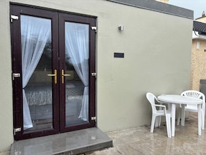 Une porte à deux battants avec des rideaux, une petite table d’extérieur avec des chaises et un luminaire fixé au mur.