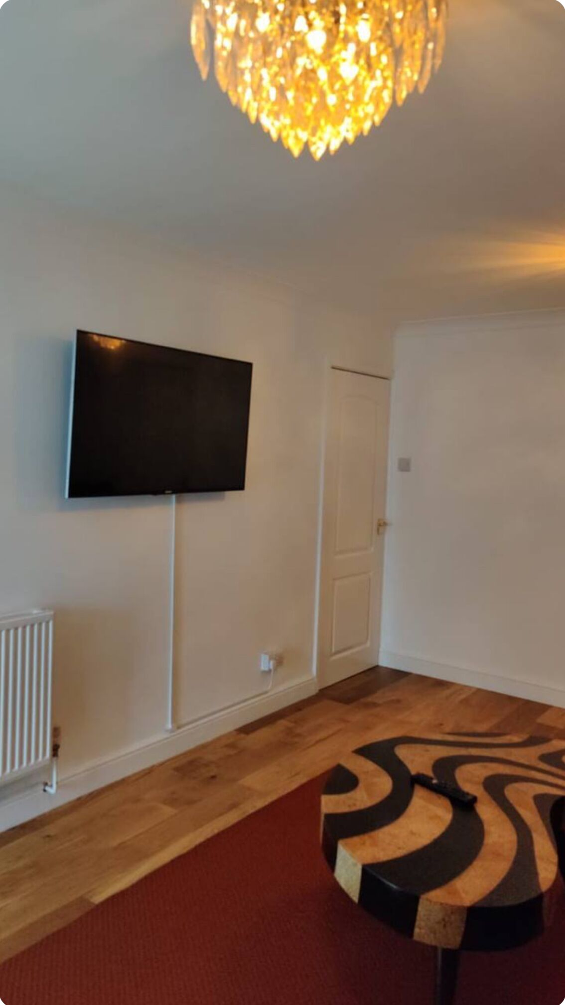 A room with a flat-screen TV mounted on the wall, a white door, a radiator, and a patterned rug on the floor.
