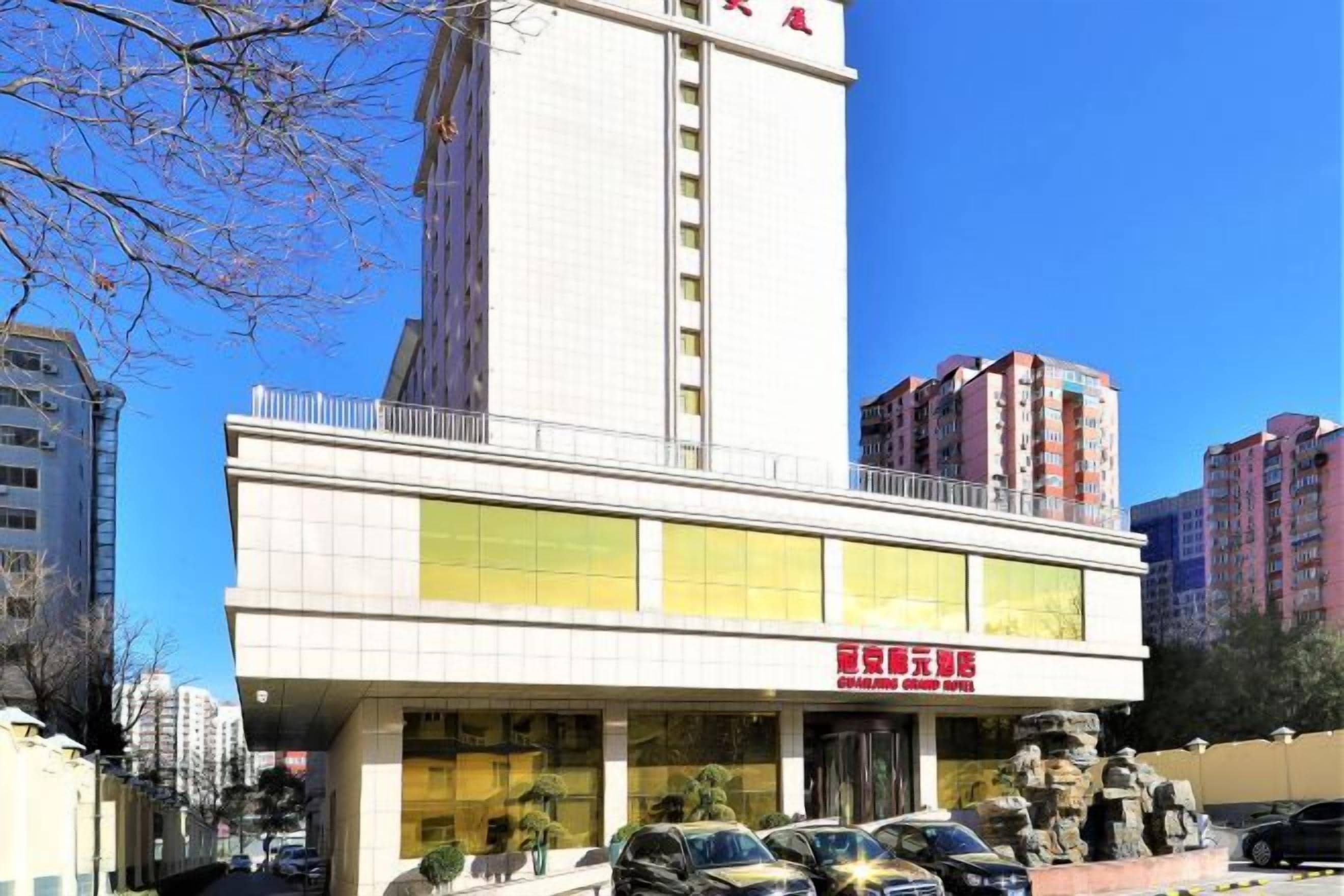 A hotel with a prominent vertical tower, a ground-level entrance with a red sign, and several parked cars in front.