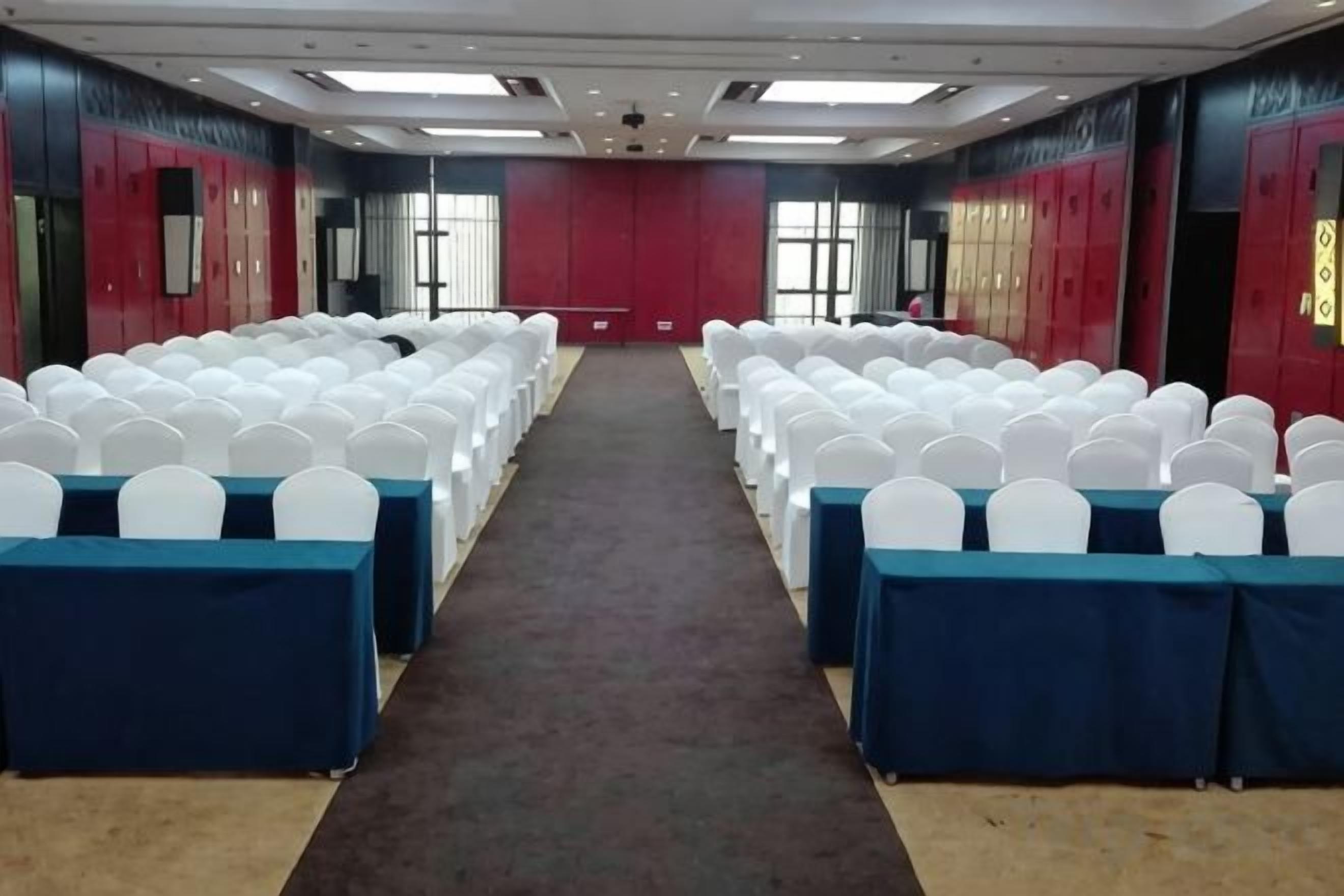 Une salle de conférence avec des rangées de chaises blanches, des nappes bleues et des murs rouges.