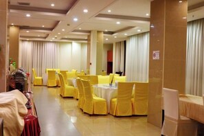 Une salle de banquet avec des tables rondes et des chaises, un espace buffet et une grande fenêtre avec des rideaux.