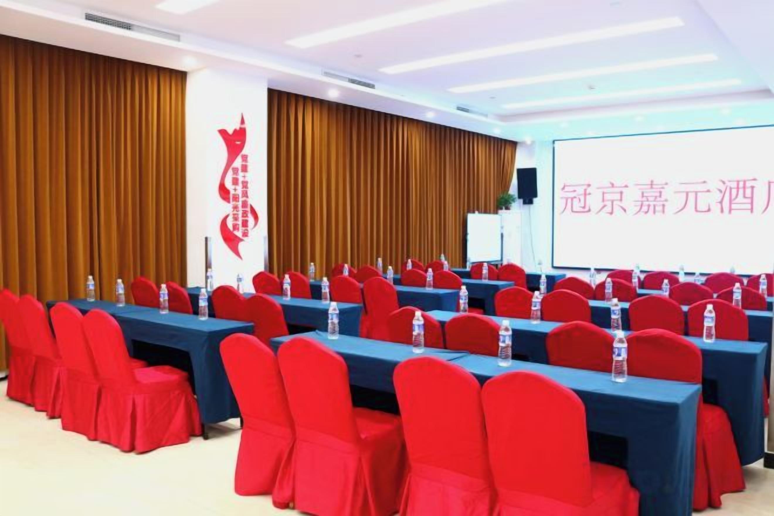 Un salón de conferencias con filas de mesas y sillas, una pantalla de proyector y un logotipo en la pared.