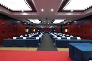Uma sala de conferências com mesas e cadeiras dispostas em fileiras, um tapete vermelho e um teto alto com luminárias.