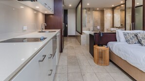 Une chambre d’hôtel moderne dotée d’une kitchenette, d’un grand lit, d’un petit tabouret en bois et d’une salle de bain avec baignoire et miroir.