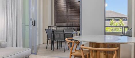 Un salon moderne avec un canapé, une table de salle à manger ronde entourée de chaises, une porte vitrée et une vue sur un bâtiment à l’extérieur.