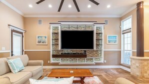 Ein Wohnzimmer mit großem Flachbildfernseher an einer Wand mit Verzierungen, beigefarbenen Sofas, einem Holztisch und einem Teppich mit Muster.