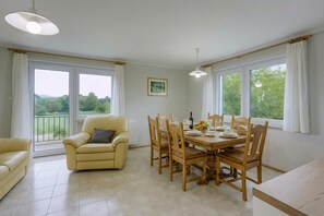 Un coin repas avec une table dressée pour quatre personnes, entouré de chaises en bois, d’un canapé et d’une fenêtre donnant sur un paysage verdoyant.