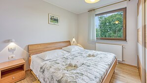 A bedroom with a wooden headboard, a large window with a view of trees, a bed with a floral quilt, and bedside lamps.